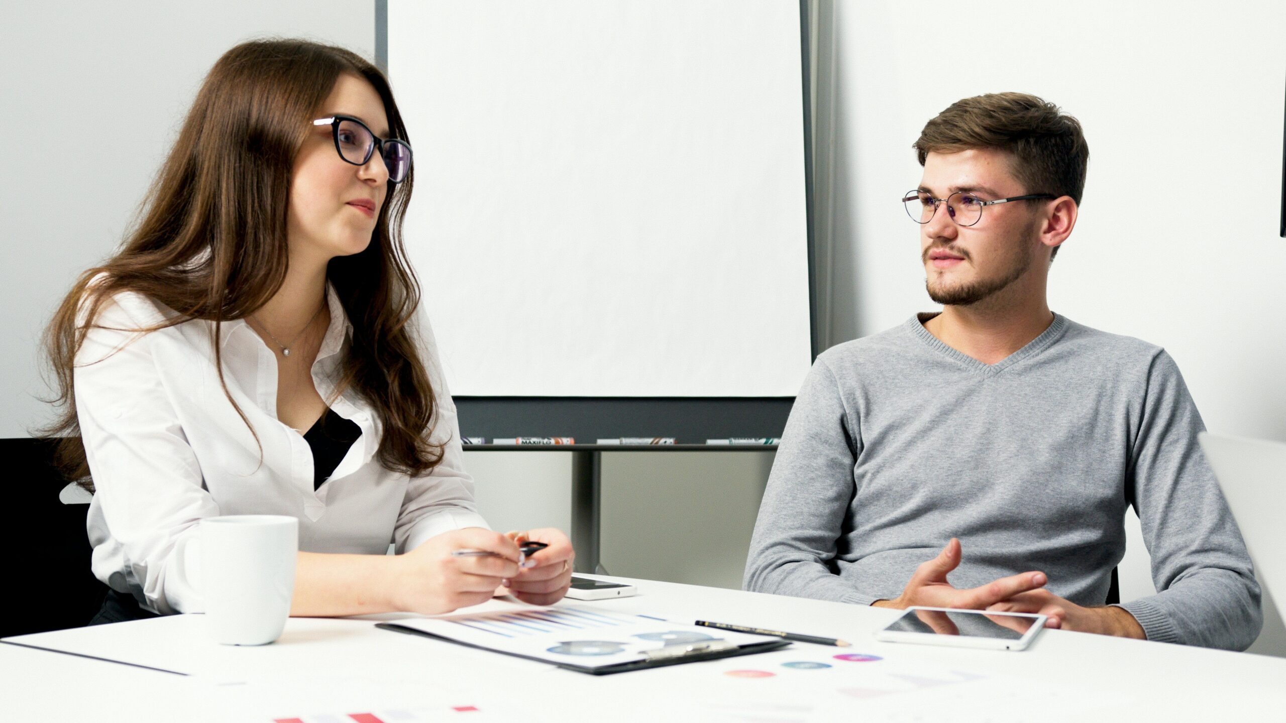 Two professionals in an office environment discussing work over a desk with documents.